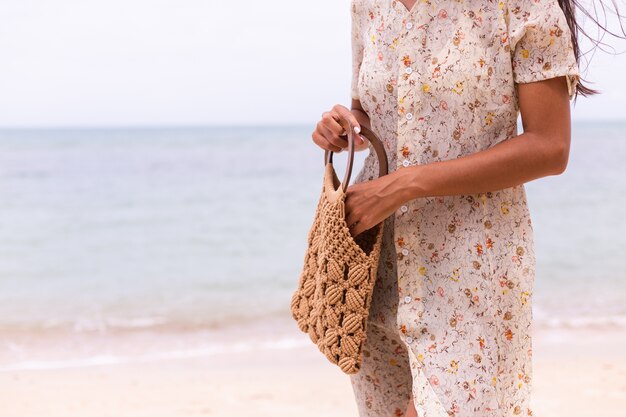 Close shot of woman in light summer flying dress holding knitted bag on beach, sea on background.