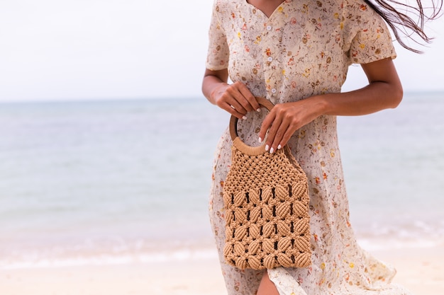 Close shot of woman in light summer flying dress holding knitted bag on beach, sea on background.