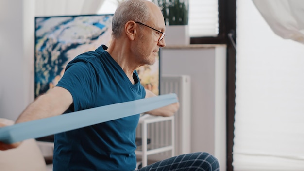 Free photo close up of aged person pulling resistance band to exercise and sitting on fitness toning ball. senior man training with elastic belt and looking at video of workout lesson on laptop