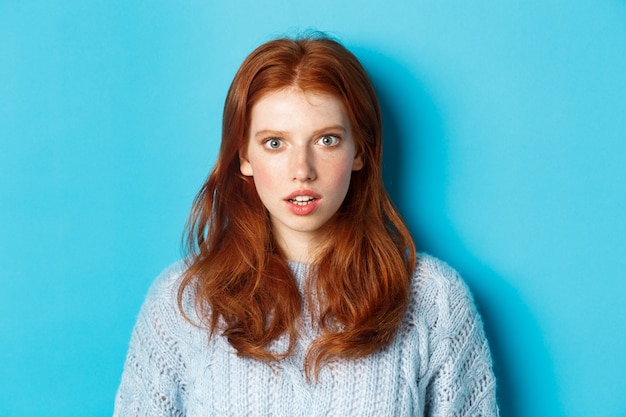 Close-up of amazed redhead girl staring at camera with complete disbelief, standing shocked against blue background