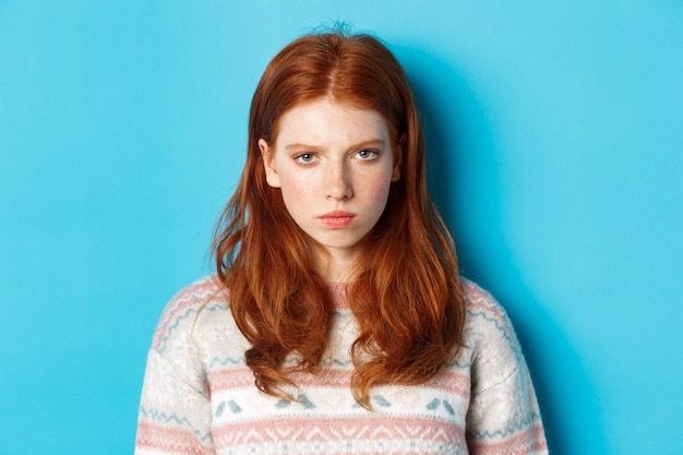 Free Photo close-up of angry redhead teenage girl looking pissed-off at camera, frowning and sulking mad, standing over blue background.