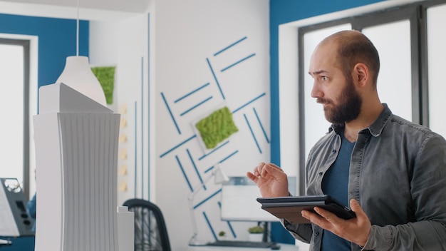 Free photo close up of architect using digital tablet and building model to work on construction design. man engineer working with device to plan property layout for architectural development project.