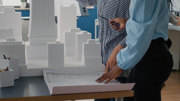 Free photo close up of architects placing blueprints plan on table to work on urban project with building model. team of women using layout print to design construction structure and development.