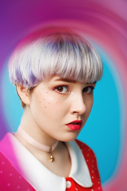 Close up artistic conceptual portrait of beautiful dollish girl with short light violet hair wearing red dress over blue wall Blurry forefront.