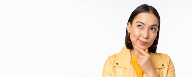 Free photo close up of asian woman thinking looking up thoughtful pondering smth with pleased smiling face standing over white background