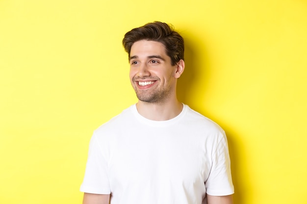 Free photo close-up of attractive bearded man in white t-shirt smiling, looking left at copy space, standing against yellow background.