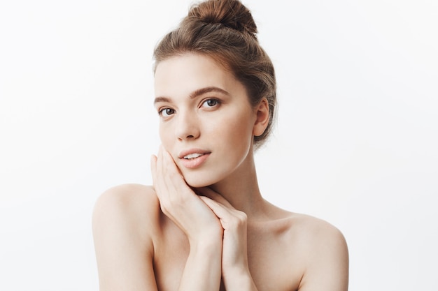 Free photo close up of attractive dark-haired young woman with bun hairstyle and naked shoulders holding hands near face,  with relaxed and calm face expression.