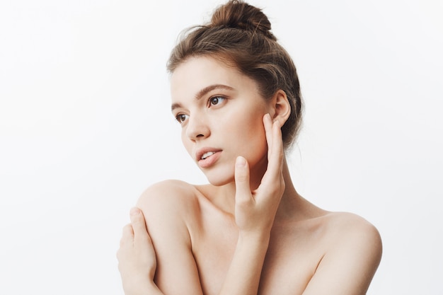 Close up of attractive tender young european woman with dark hair in bun hairstyle being naked looking aside with calm expression, touching face with hands posing for magazine photo shoot.