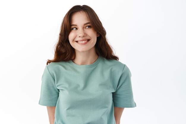 Close up of attractive young brunette woman looking aside at your promotional text staring at banner with pleased happy smile standing against white background