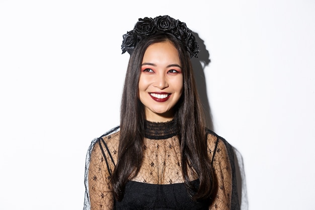 Free photo close-up of beautiful elegant woman in witch costume and gothic makeup, smiling pleased while looking at upper left corner, celebrating halloween.