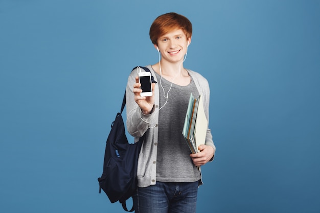 Free photo close up of beautiful ginger student boy with backpack smiling, holding lot of notebooks in hands, showing cell phone, listening to favorite music in earphones.