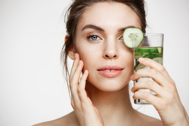 Free Photo close up of beautiful nude girl smiling looking at camera holding glass of water with cucumber slices over white background. healthy nutrition. beauty and skincare.