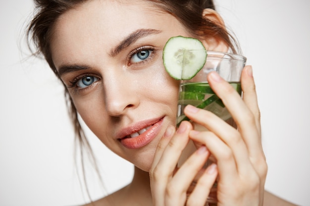 Free Photo close up of beautiful nude girl smiling looking at camera holding glass of water with cucumber slices over white background. healthy nutrition. beauty and skincare.