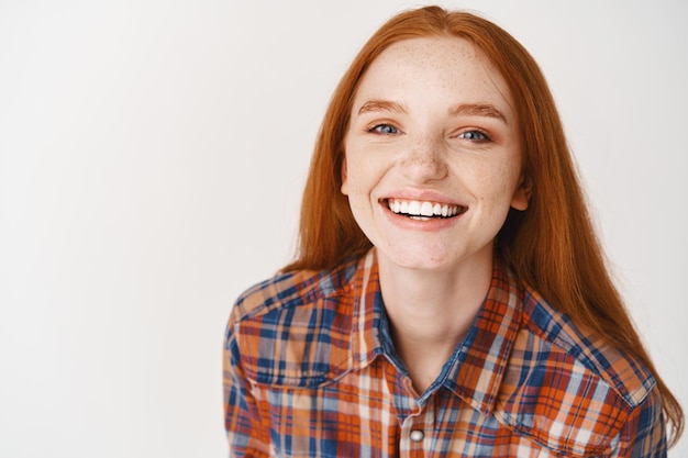 Close-up of beautiful redhead female model with pale skin and white teeth, smiling happy at front, standing over white wall