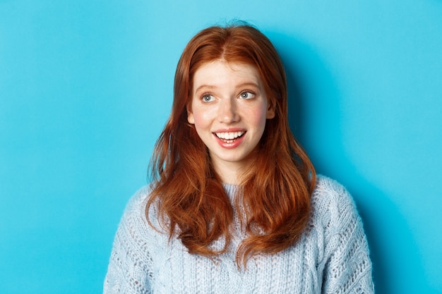 Close-up of beautiful redhead girl in sweater, looking at upper left corner promo logo and smiling, standing over blue background.