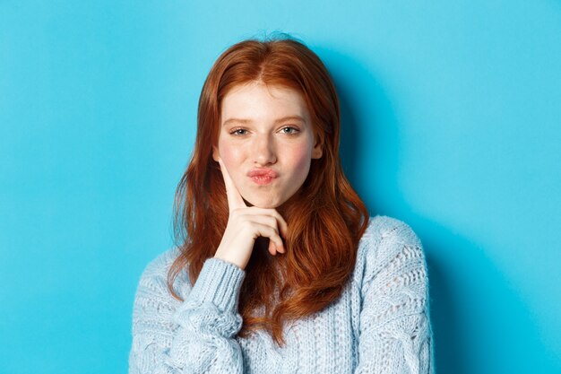 Close-up of beautiful redhead girl thinking, pucker lips and staring thoughtfula t camera, making choice, standing over blue background.