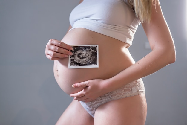 Close-up of belly of young pregnant model showing ultrasonic image of her baby. Future mom on her second trimester holding ultrasound scan of her child. Maternity concept