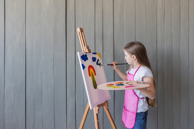 Free photo close-up of a blonde girl painting with paint brush on easel standing against gray wooden wall