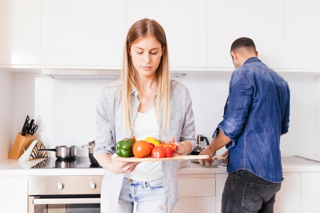 Close-up of blonde young woman holding colorful bell peppers in hand with her husband at background