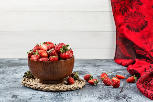 Free Photo close-up a bowl of strawberries on round wicker placemat with red scarf on dark blue marble and white wooden board surface. horizontal free space for your text