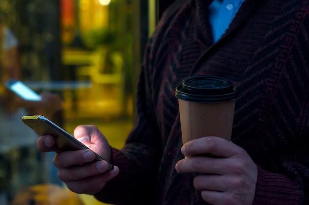 Free photo close up of a businessman using mobile phone and holding paper cup. close-up detail of a businessmans hand holding paper cup and using a smartphone while walking on the road.