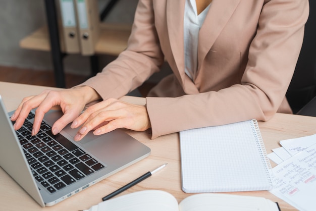 Close-up of businesswoman's hand typing on laptop with pen; diary and spiral notepad on wooden table