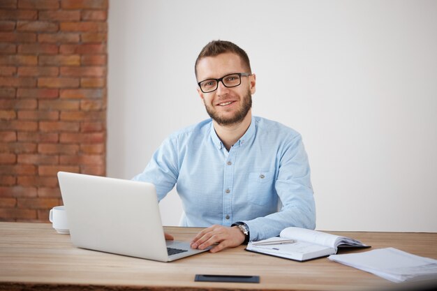 Close up of cheerful adult bearded male manager in glasses and classic shirt sitting at desk in office, working at personal computer, writing information in notebook.