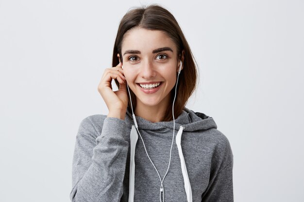 Close up of cheerful young attractive dark-haired caucasian girl in casual gray hoodie smiling brightly, holding earphone with hand,  with happy and relaxed face expression.