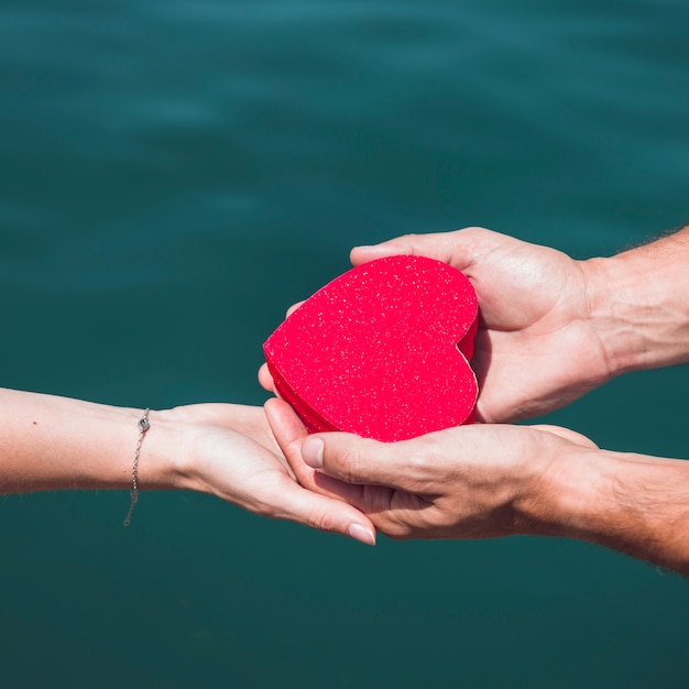 Free photo close-up of a couple's hand holding red heart shape in front of sea