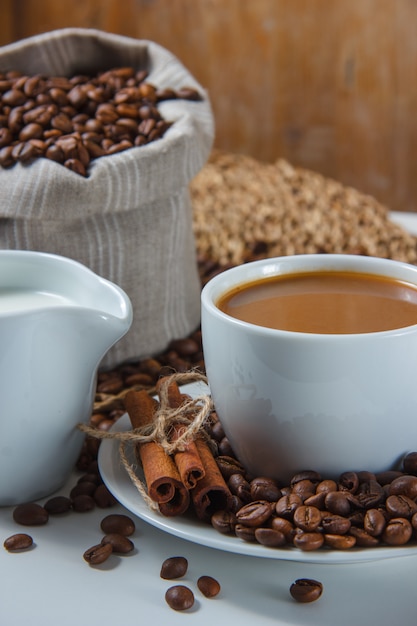 Close-up a cup of coffee with coffee beans in a sack and saucer, milk, dry cinnamon on trivet and white surface. vertical