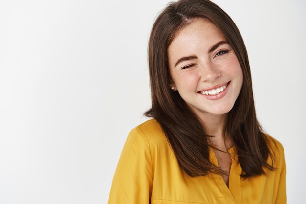 Close-up of cute and dreamy young woman smiling, winking at front to encourage client, standing over white wall