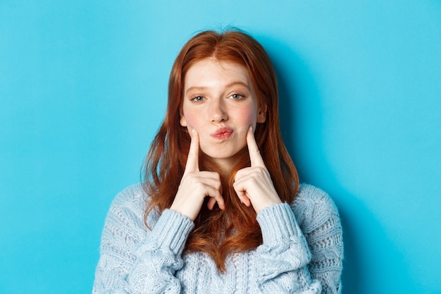 Close-up of cute redhead girl in sweater, pucker lips and pointing fingers at cheeks, poking dimples, standing over blue background