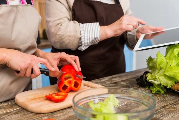 Free Photo close-up of daughter standing with her mother holding digital tablet cutting bell pepper with knife