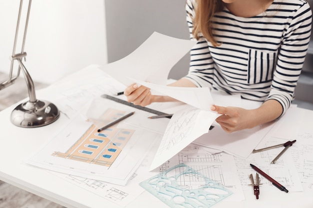 Close up detail of female engineer wearing striped clothes sitting at white big table, looking through papers, working on construction project.