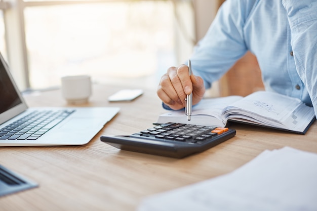 Close up detail of professional serious accountant sitting in light office, checking company finance profits on calculator