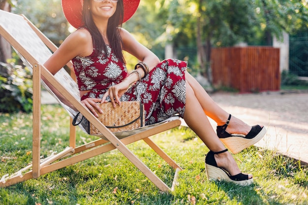 Close up details legs wearing wedges sandal shoes, footwear, stylish beautiful woman sitting in deck chair in tropical style outfit, summer fashion trend, straw handbag, accessories, vacation