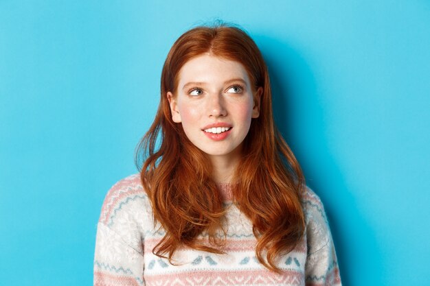 Close-up of dreamy redhead girl imaging something, staring at upper right corner and smiling, standing in winter sweater against blue background