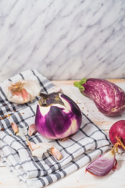 Close-up of eggplants; onion; garlic cloves and chequered pattern textile on wooden surface