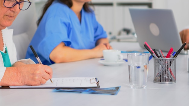 Free Photo close up of elderly woman doctor taking notes on clipboard while coworkers discussing in background, writing on laptop during brainstorming. profesional teamworker having medical meeting