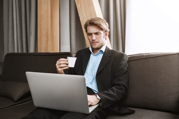 Free photo close up of elegant unshaven man drinking coffee, looking in laptop monitor with serious and unsatisfied expression, working from home.