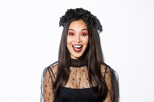 Close-up of enthusiastic asian woman looking amused, wearing witch costume on halloween, standing over white background.