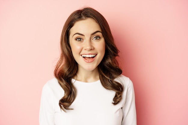 Free photo close up of enthusiastic brunette girl with makeup, smiling and looking happy at camera, posing against pink background.