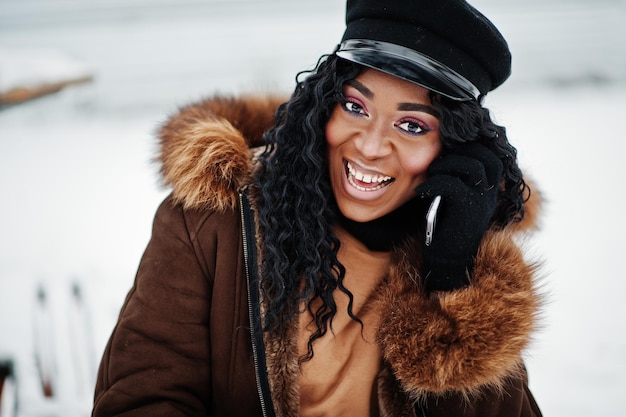 Close up face of african american woman in sheepskin coat and cap posed at winter day against snowy background with phone at hand