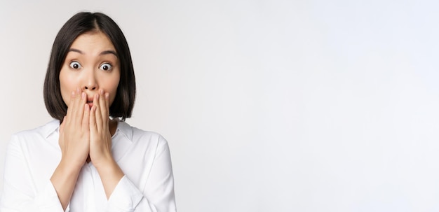 Close up face of asian woman gasping looking shocked and speechless holding hands near mouth and staring at camera impressed white background