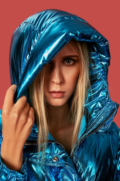 Free photo close up fashion photo of young woman covering herself with hood. she wears blue oversized down jacket in studio on red background