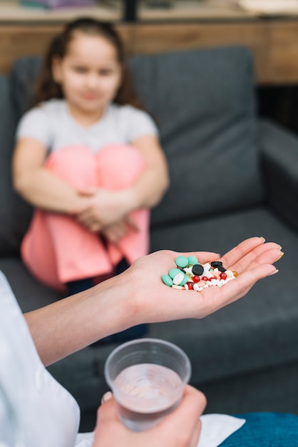 Close-up of female doctor's hand with different pills in front of a girl sitting on sofa