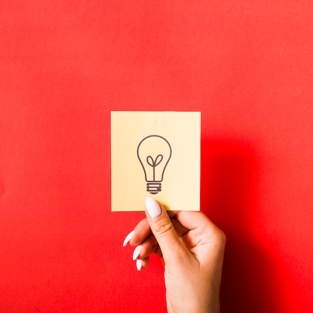 Free photo close-up of female hand holding sticky note with drawn light bulb on red background