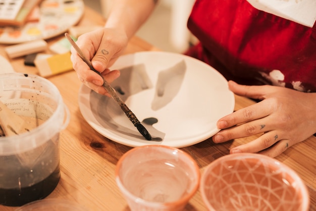 Close-up of female painting the tableware with paintbrush on wooden table