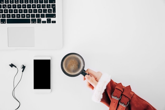 Close-up of female's hand holding coffee cup over the office desk with laptop; mobilephone and earphone