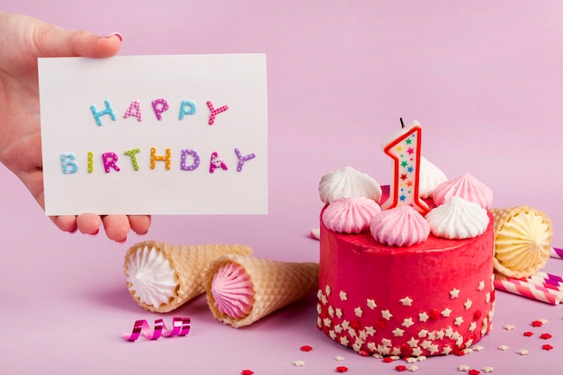 Close-up of a female's hand holding happy birthday card near the decorative cake against purple backdrop